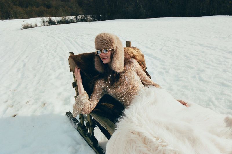 Woman in fur hat and sequin dress reclines on fur-covered sled in snowy landscape. Trees visible in background, sunny day.