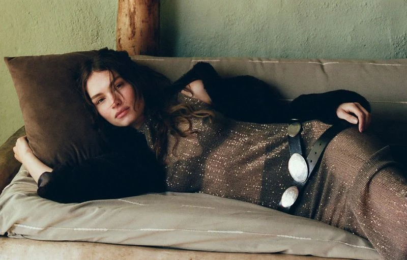 Adult woman poses lying on a sofa with a pillow, wearing a dress with sequins and a black jacket, indoors with a minimalist aesthetic.