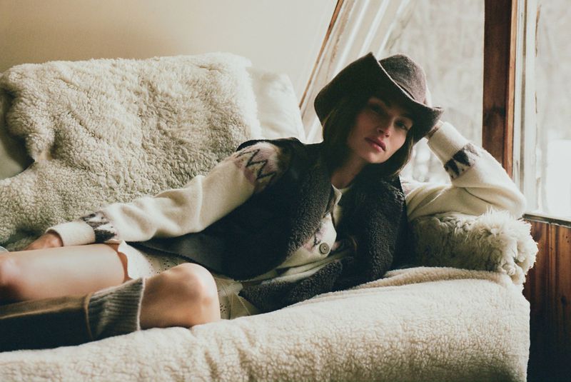 A woman in a hat and vest is lounging on a textured sofa near a window, illuminated by natural light in an indoor setting.