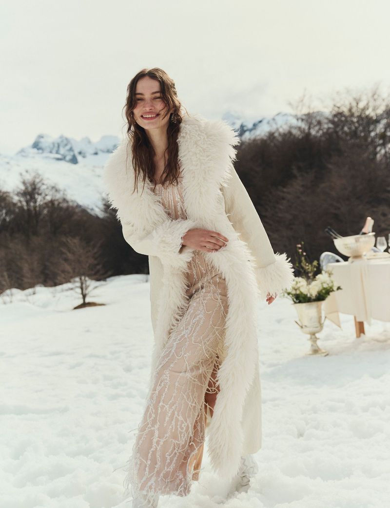 Smiling woman in a fur-trimmed coat poses in a snowy mountain landscape with a party table, exuding elegance and winter charm.
