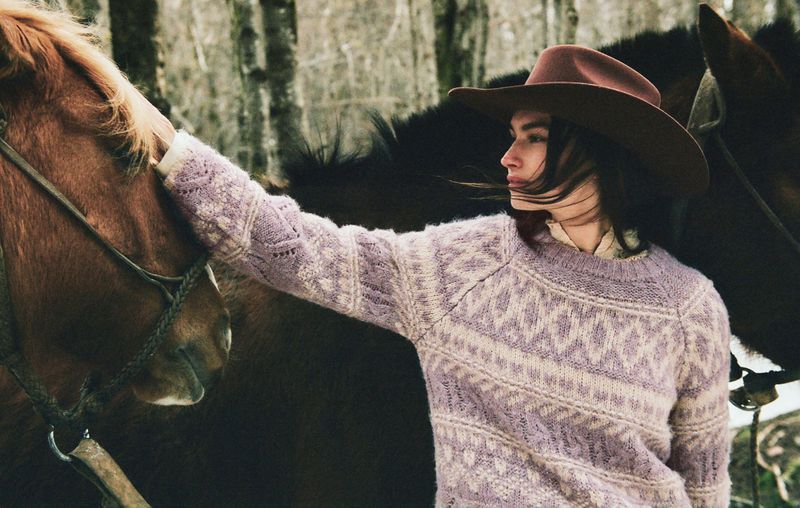 Woman in lilac sweater and cowboy hat stands next to brown horses in wintry scene, shallow depth of field, muted colors, and soft focus.