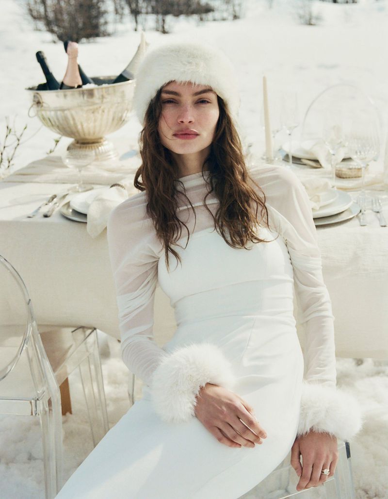 Woman in a white dress and fur hat poses in a snowy setting near a table with champagne. Fashion shot, winter scene, outdoor setting.