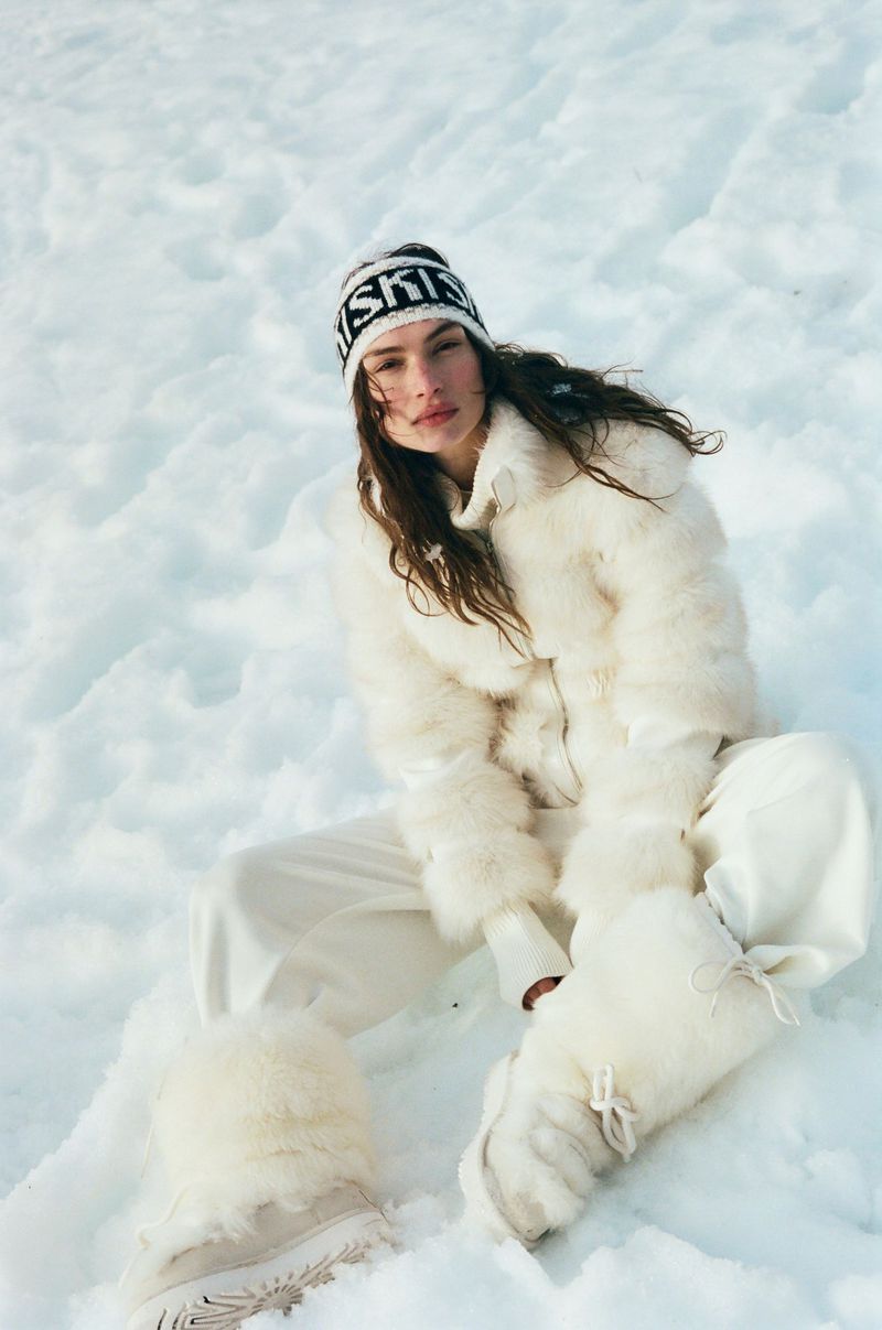 Fashionable young adult female in a white fur jacket, ski hat, and boots sits in the snow against a winter background.