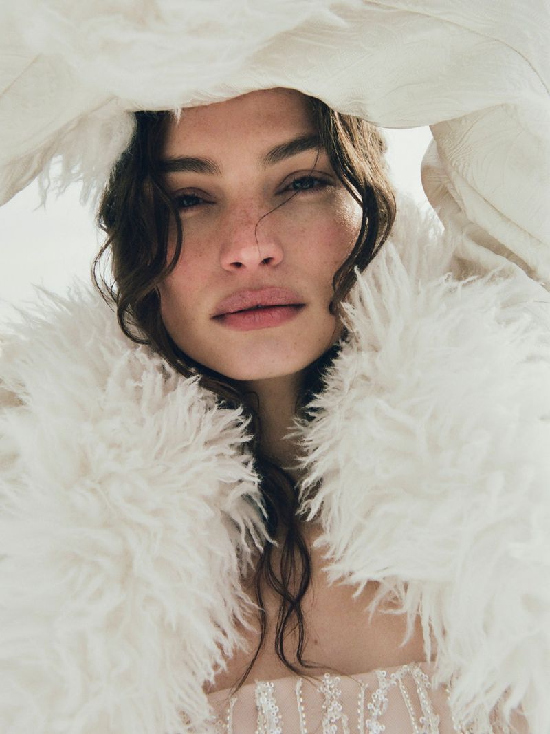 Close-up portrait of an adult woman with wavy dark hair and fair skin wearing a feathered garment against a bright background.