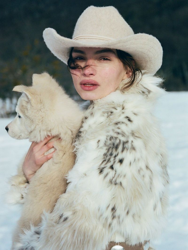 Close-up of woman in cowboy hat and spotted fur coat holding a white husky puppy in a snowy, blurred outdoor setting.