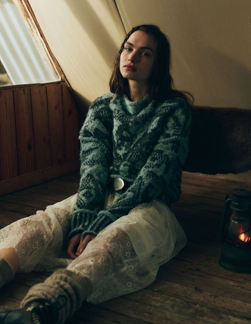 Woman poses indoors on wooden floor in green patterned sweater, lace pants, and leg warmers by a window and lit lantern.