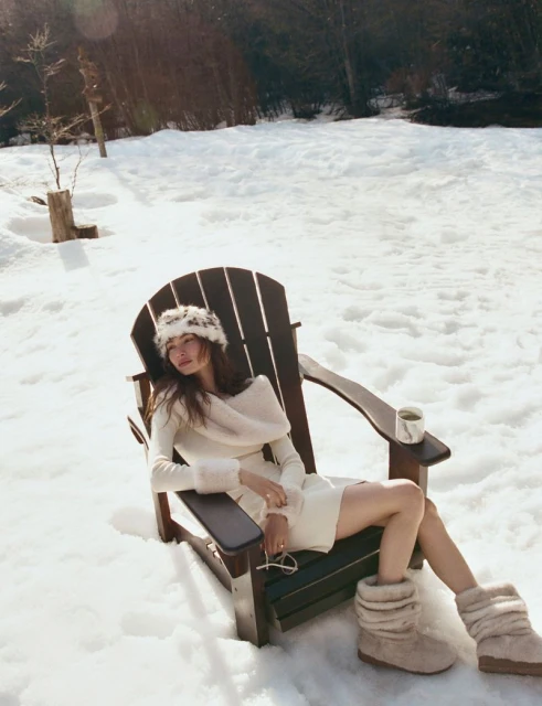 Woman in cream dress and fuzzy hat sits in snow in an Adirondack chair in a winter landscape, wearing fuzzy boots and smiling.