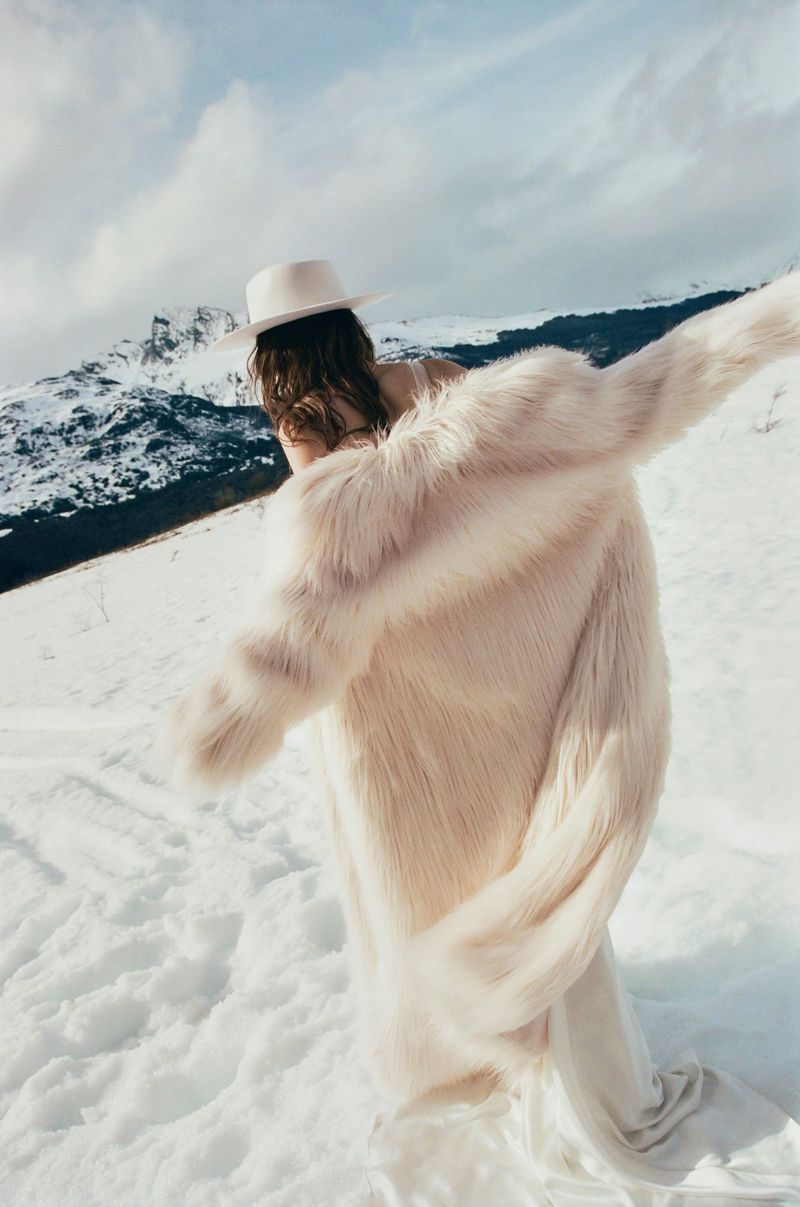 A woman in a fur coat and hat stands in the snow in a mountain landscape with arms outstretched, creating a fashionable outdoor portrait.