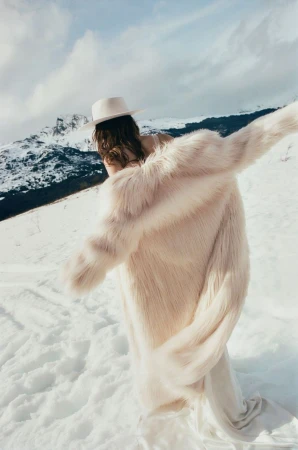 Model wearing a white fedora hat and fluffy faux fur coat walking across a snowy mountain landscape