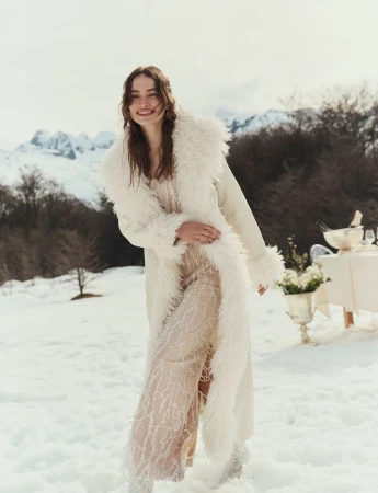 Model Valerie Pasker smiling in the snow wearing a long cream coat with shaggy fur trim and a sheer embroidered dress.