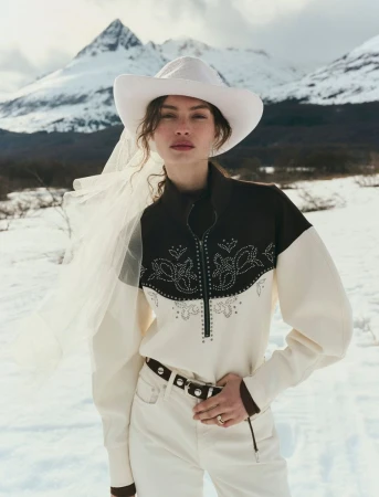 Model wearing a white cowboy hat with a veil and brown-and-white patterned jacket against a snowy mountain backdrop.