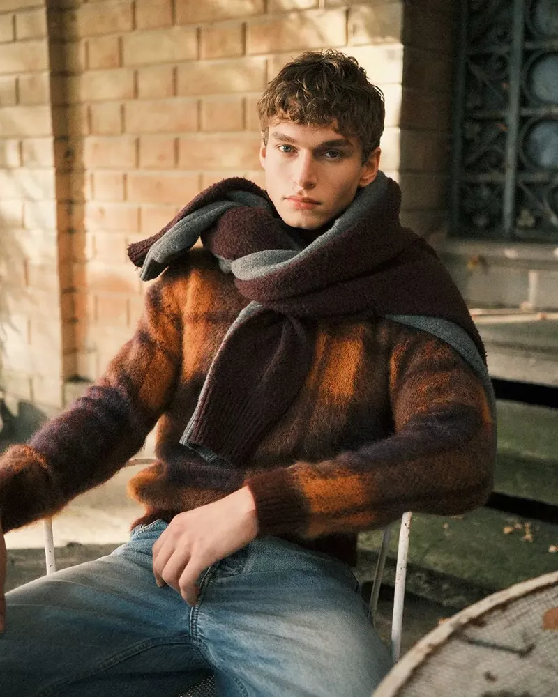 A young adult male model is sitting outside wearing a brown and orange sweater and grey scarf with a neutral expression on his face.