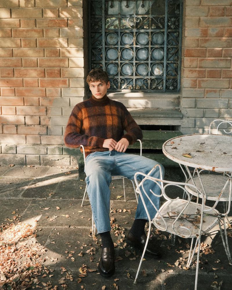 A young man in a patterned sweater and jeans sitting outside on a vintage metal chair and table with a brick building backdrop.