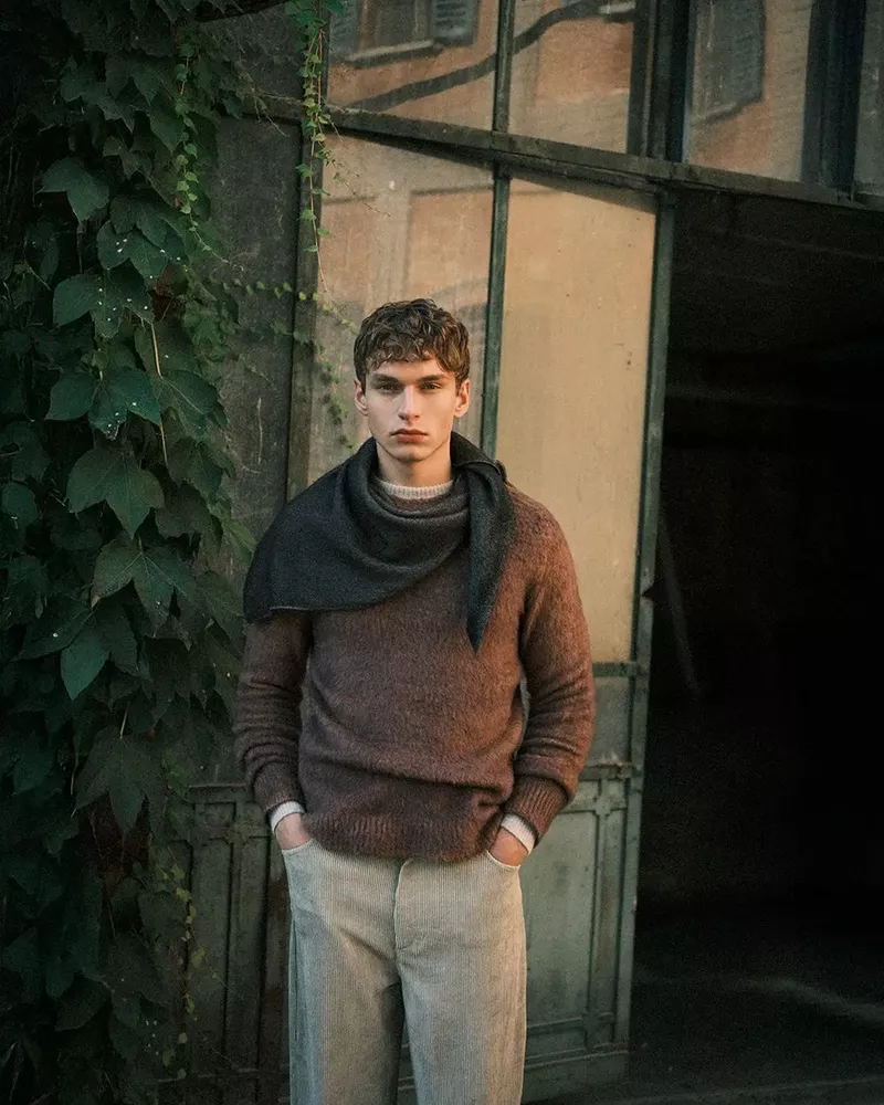 Adult man in casual brown sweater and neutral pants posing outdoors near a vine-covered architectural structure.