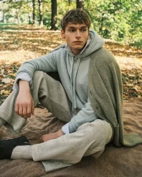Jan Rykowski sits on a blanket among fallen leaves wearing a light textured hoodie, sage green knit cardigan, and ribbed trousers.