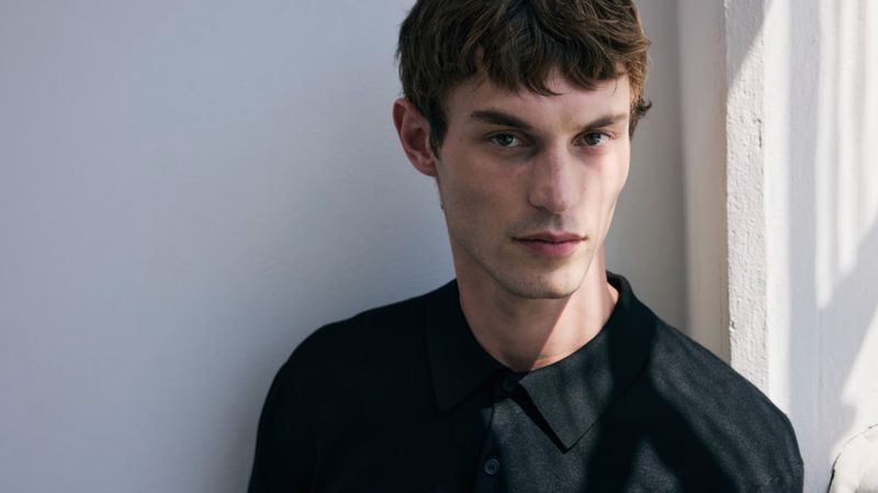 Close-up studio portrait of a young adult male with dark brown hair wearing a black collared shirt, posing against a bright white wall.