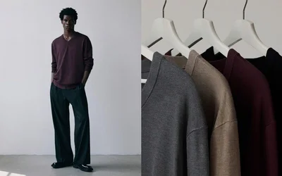 A male model in a burgundy v-neck sweater and wide-leg trousers standing beside a rack of hanging knit sweaters on white hangers.