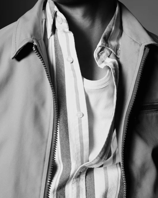 A close-up photograph shows an open light-brown jacket revealing a striped button-down shirt and plain white t-shirt underneath. The textures are soft.