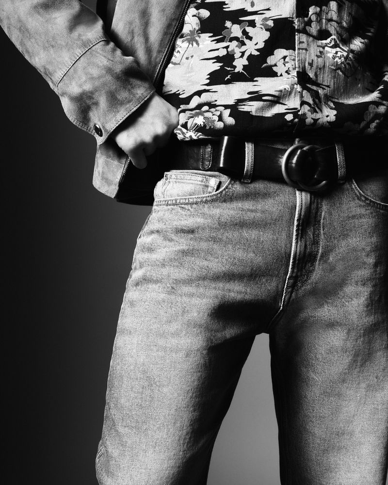Monochromatic studio shot of a person wearing jeans, a floral shirt, and a belt with a round buckle. Jacket is visible on the shoulders.