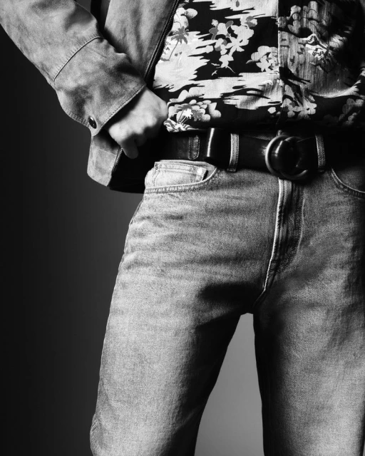 Monochromatic studio shot of a person wearing jeans, a floral shirt, and a belt with a round buckle. Jacket is visible on the shoulders.