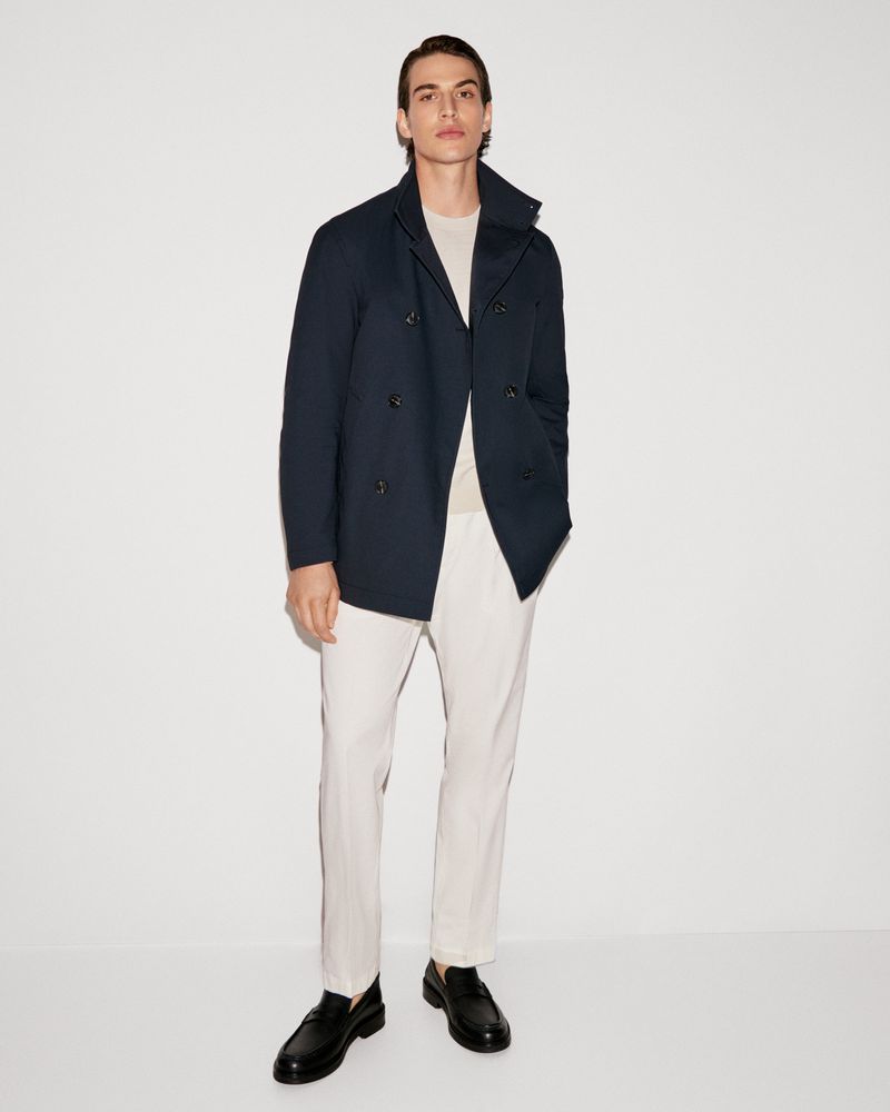A young adult male posing in a navy peacoat, light beige pants, and black loafers against a white background, shot in portrait orientation.