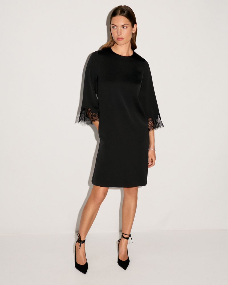 Elegant adult female model poses in a black short dress with lace sleeves and ankle-tie heels against a stark white backdrop.