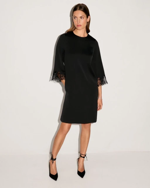 Elegant adult female model poses in a black short dress with lace sleeves and ankle-tie heels against a stark white backdrop.