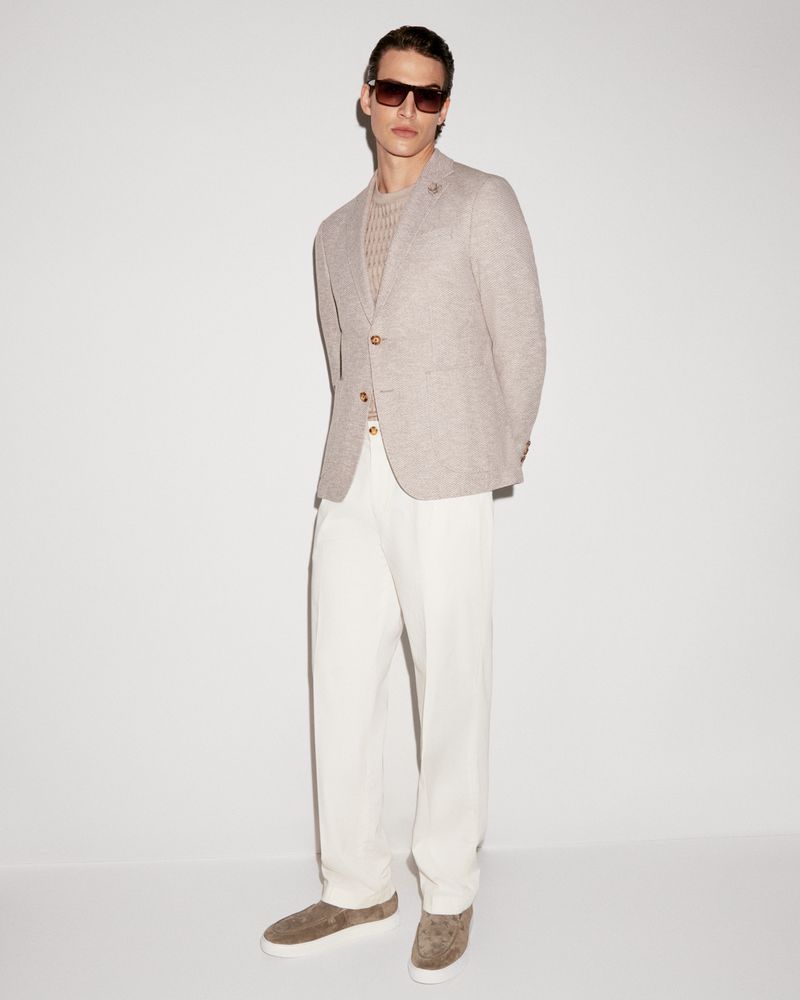 Man stands in front of a white wall wearing sunglasses, a tan blazer, light beige top, cream pants, and patterned sneakers.