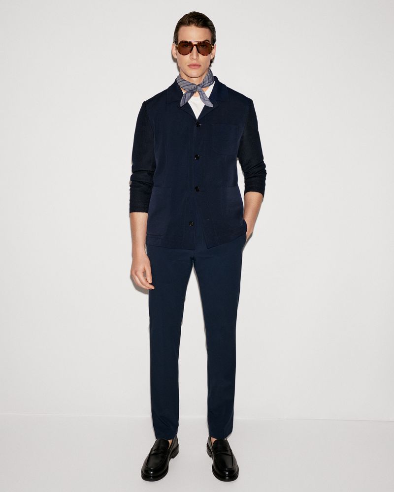 A man in a navy suit, white shirt, patterned neck scarf, sunglasses and loafers, stands against a plain white backdrop in a studio.
