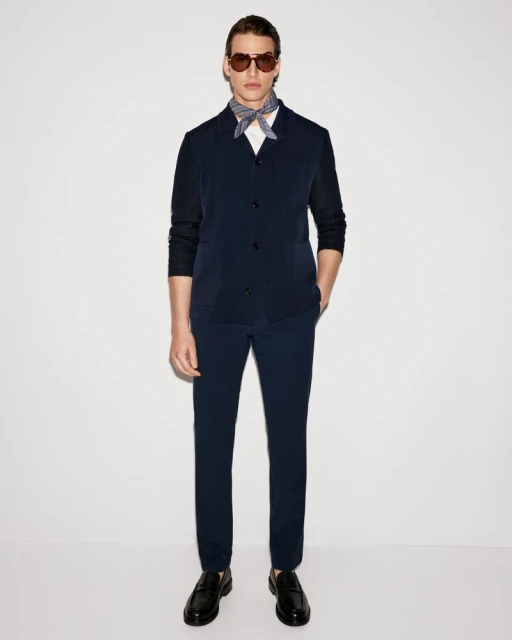 A man in a navy suit, white shirt, patterned neck scarf, sunglasses and loafers, stands against a plain white backdrop in a studio.