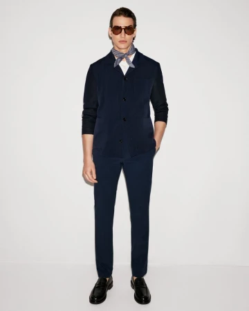 A man in a navy suit, white shirt, patterned neck scarf, sunglasses and loafers, stands against a plain white backdrop in a studio.