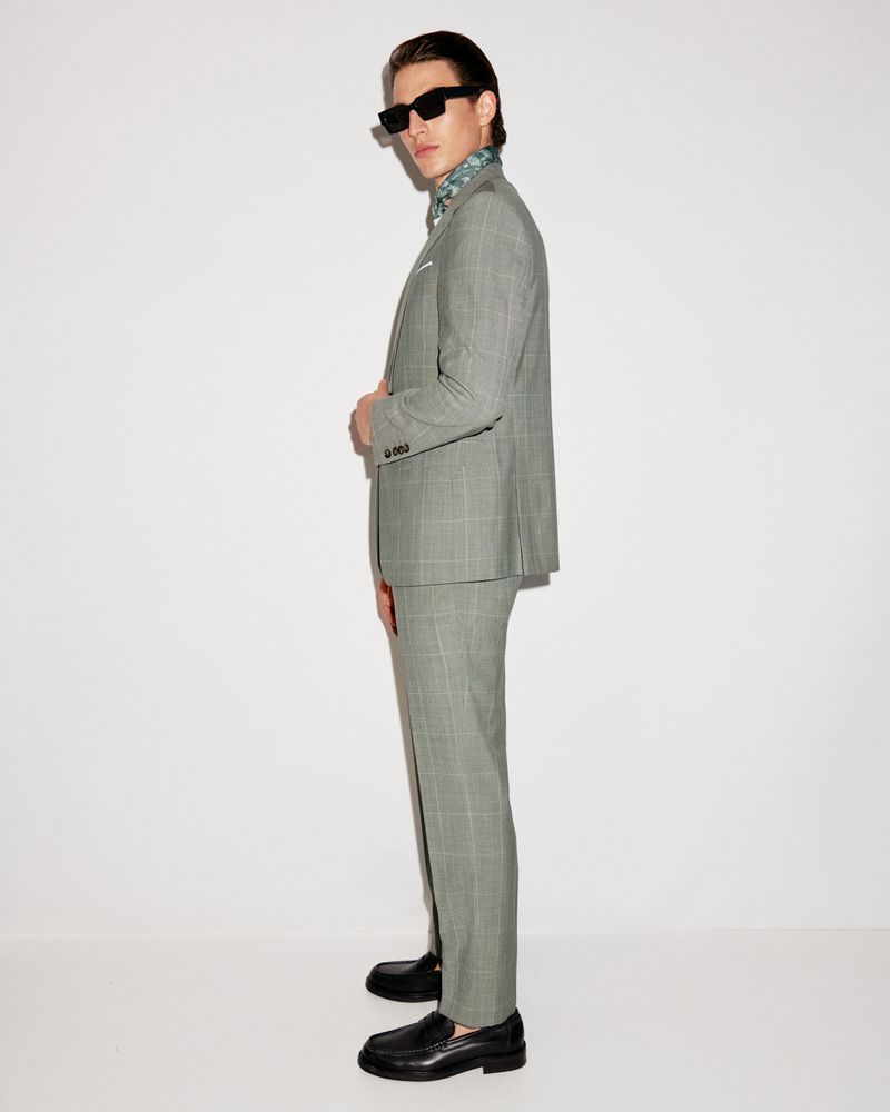 Man in grey plaid suit with black shoes, posing in front of a white wall. He is wearing dark glasses and a patterned ascot.