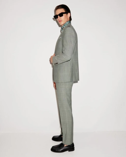 Man in grey plaid suit with black shoes, posing in front of a white wall. He is wearing dark glasses and a patterned ascot.