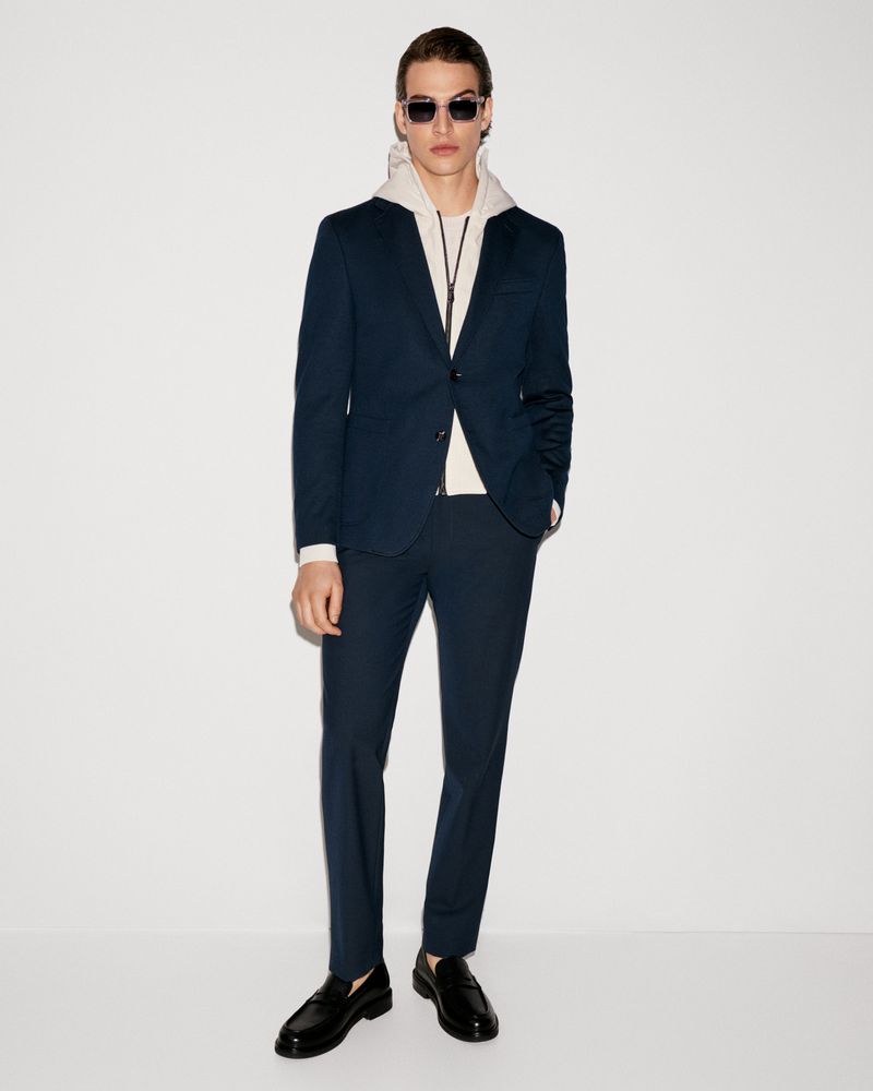 Male fashion model posing in a navy suit with a cream-colored hoodie and black loafers, standing against a clean white background.