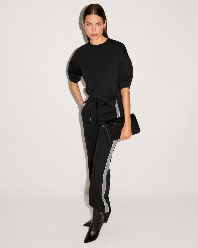 Elegant woman in black sweater and track pants poses with black clutch bag and heeled boots in a neutral photo studio backdrop.