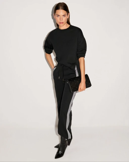 Elegant woman in black sweater and track pants poses with black clutch bag and heeled boots in a neutral photo studio backdrop.