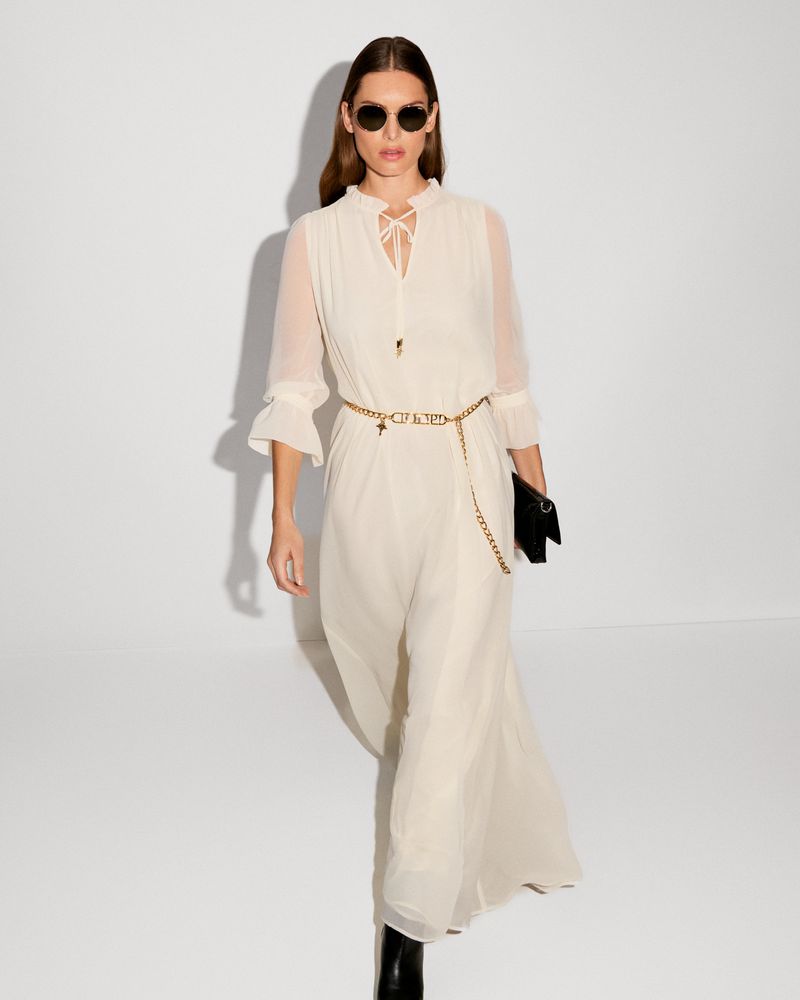 A woman in a flowing white dress, accessorized with sunglasses and a chain belt, strides against a bright white studio backdrop.