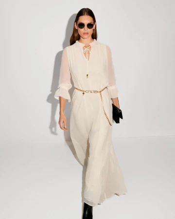 A woman in a flowing white dress, accessorized with sunglasses and a chain belt, strides against a bright white studio backdrop.