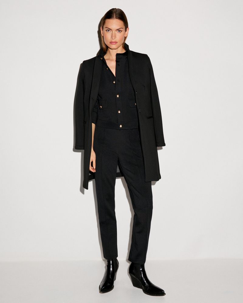 Model wearing a black coat and matching trousers against a plain white studio backdrop.