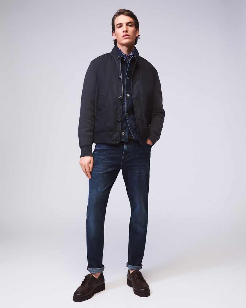 Full-length shot of an adult male wearing a black jacket, blue jeans, and brown shoes, posing in a photo studio with a neutral background.