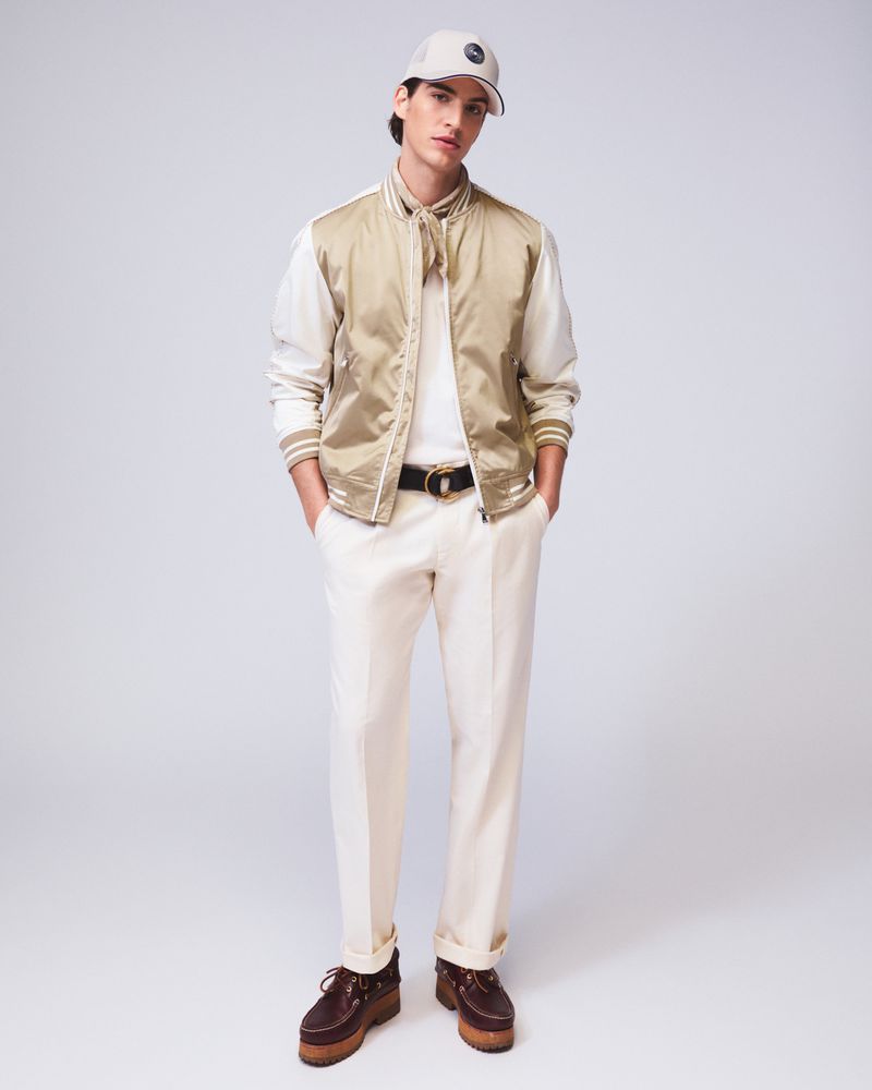 Full-shot of a young adult man posing in a tan jacket, cream trousers, dark belt, and cap against a neutral grey backdrop.