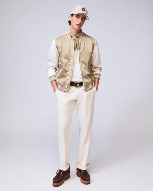 Full-shot of a young adult man posing in a tan jacket, cream trousers, dark belt, and cap against a neutral grey backdrop.