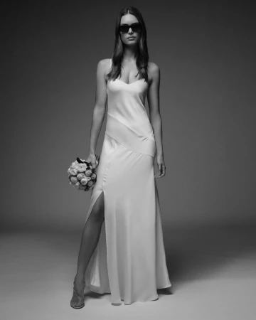 Model wearing dark sunglasses and a long white satin gown with a high slit, holding a small floral bouquet against a studio backdrop.