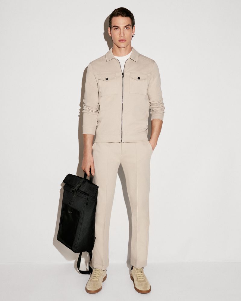 Full-length studio shot of a male model wearing a neutral-colored suit, sneakers, and holding a black backpack against a white backdrop.
