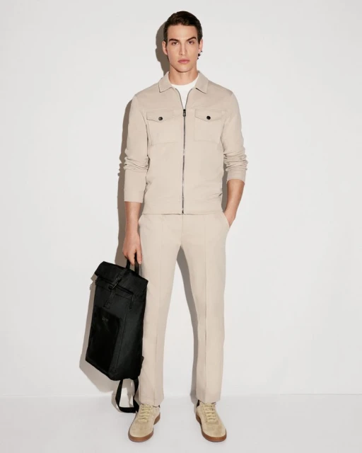 Full-length studio shot of a male model wearing a neutral-colored suit, sneakers, and holding a black backpack against a white backdrop.