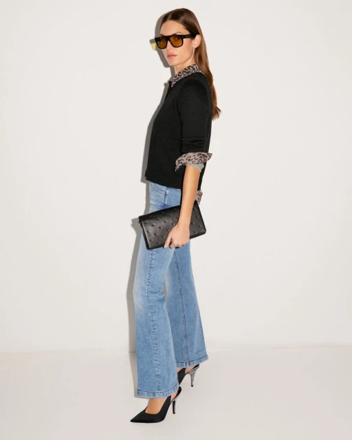 Full shot of a woman posing in sunglasses, black sweater, blue jeans, and black heels in a photo studio against a white backdrop.