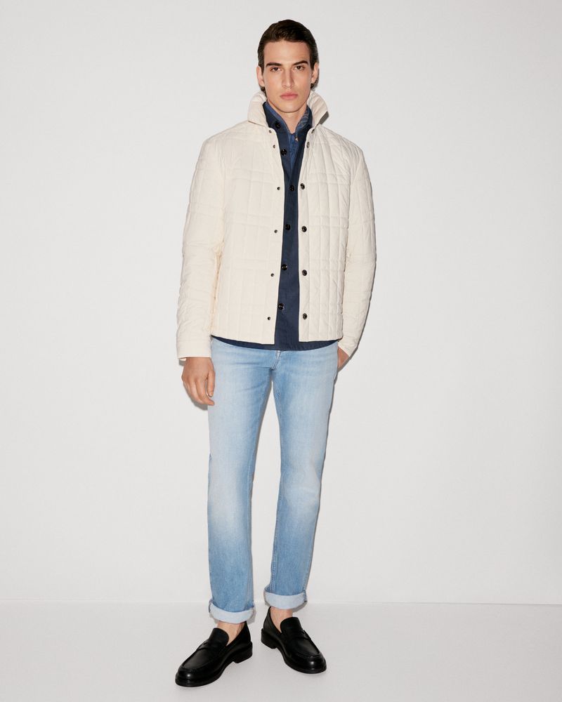 Full-length shot of a young adult man in casual fashion, wearing a quilted jacket, button-down shirt, light-wash jeans, and loafers against a white backdrop.