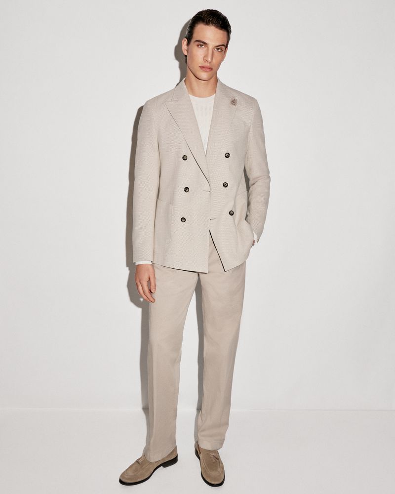A man stands in a light tan double-breasted suit, paired with matching pants and suede loafers, set against a bright white background.