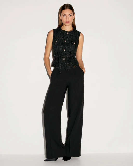 A woman stands in a photo studio wearing a black and metallic top and black slacks, posing with her hands in her pockets.