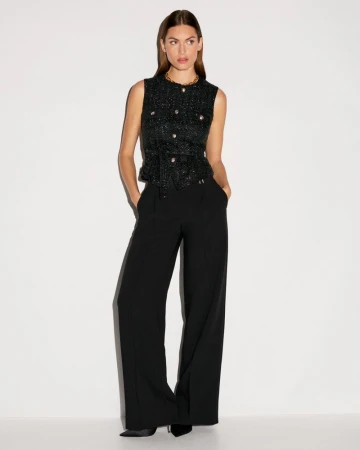 A woman stands in a photo studio wearing a black and metallic top and black slacks, posing with her hands in her pockets.